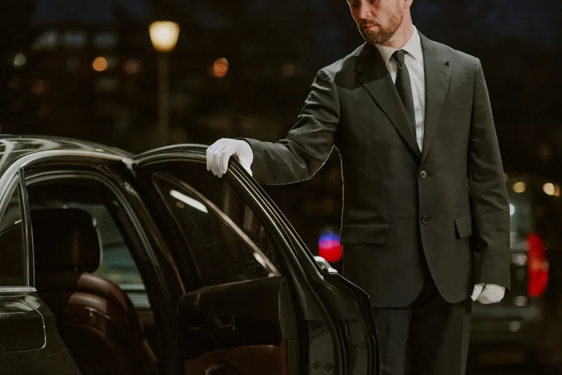 A suited chauffeur holding open the door of a black car for a waiting passenger
