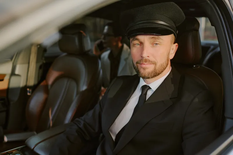 A professional chauffeur in a suit and cap at the wheel of a luxury car